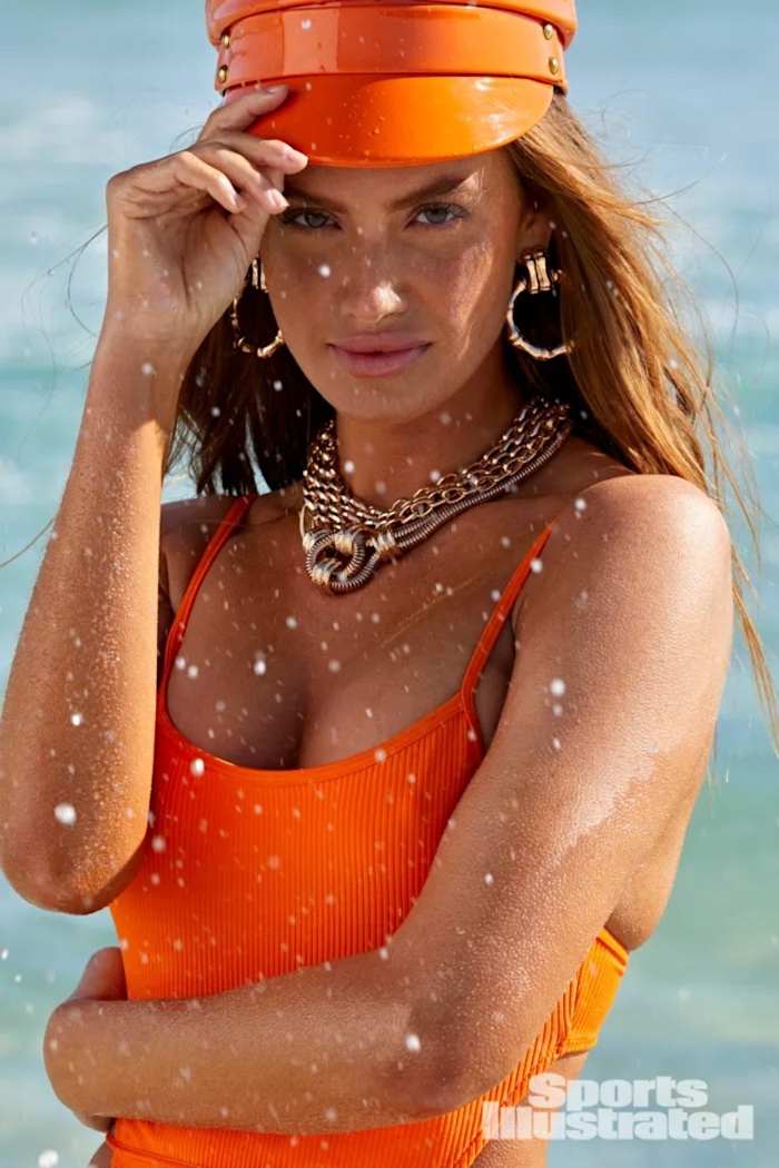 Haley Kalil stands in the ocean in a bright orange one-piece, orange visor and oversized gold jewelry.